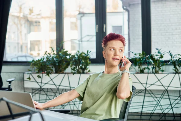 Bigender uomo d'affari che parla sul cellulare vicino al computer portatile e distogliendo lo sguardo in ufficio moderno — Foto stock