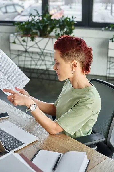 Vista laterale di elegante persona non binaria che lavora con documenti vicino laptop e telefono cellulare in ufficio — Foto stock