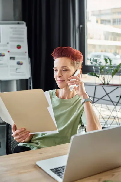 Persona queer ocupado hablando en el teléfono inteligente y mirando los documentos cerca de la computadora portátil en la oficina moderna - foto de stock