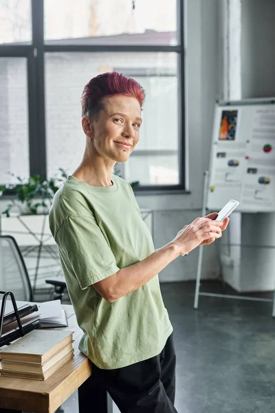 Gaie gestionnaire queer réussie avec téléphone mobile regardant la caméra tout en restant debout dans le bureau — Photo de stock