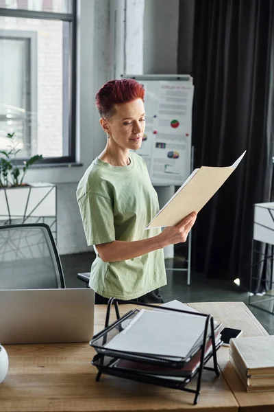 Elegante queer manager segurando pasta com documentos e de pé perto laptop na mesa de trabalho no escritório — Fotografia de Stock