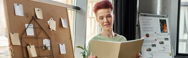 Persona queer sonriente mirando los documentos cerca de corcho con notas de papel en la oficina, pancarta - foto de stock