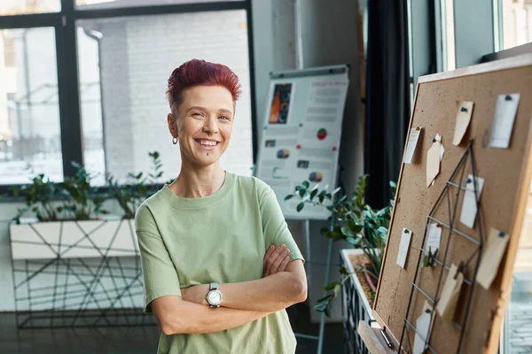 Happy non-binary person with short hair looking at camera near corkboard with paper notes in office — Stock Photo