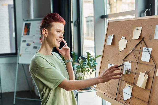 Stylish queer person talking on mobile phone and looking at paper notes on corkboard in office — Stock Photo