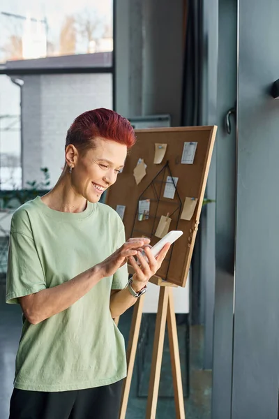 Joyful short haired bigender manager in casual attire messaging on smartphone in contemporary office — Stock Photo