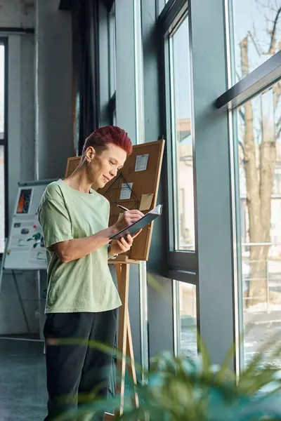 Elegante administrador queer escribir en el cuaderno, mientras que de pie cerca de las ventanas en el entorno moderno de la oficina - foto de stock