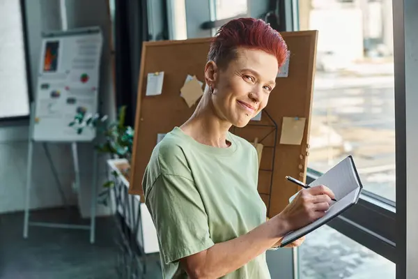 Elegante manager queer sorridente alla fotocamera e scrivere in quaderni in ambiente ufficio moderno — Foto stock