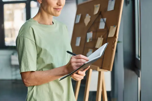 Cropped view of stylish non-binary manager standing in modern office and writing task in notebook — Stock Photo