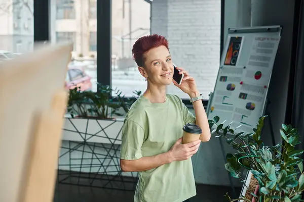 Happy queer person with coffee to go in paper cup talking on mobile phone in modern office — Stock Photo
