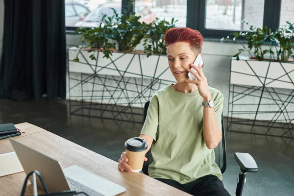 Позитивна квітка людина з кавою, щоб піти в паперову чашку, розмовляючи на мобільному телефоні біля ноутбука в офісі — стокове фото