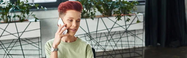 Positive queer person with short hair talking on mobile phone in modern office, horizontal banner — Stock Photo