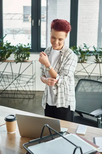 Fröhliche Bigender-Managerin in lässiger Kleidung, die am Arbeitsplatz steht und im Büro auf Laptop schaut — Stockfoto