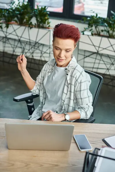 Elegante persona no binaria con pluma sentada en el lugar de trabajo cerca de la computadora portátil y el teléfono inteligente con pantalla en blanco - foto de stock