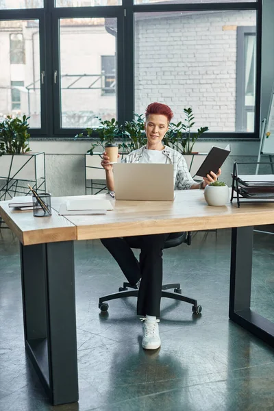 Стильна небінарна людина з кавою, щоб піти і блокнот, що сидить біля ноутбука в сучасному офісі — стокове фото
