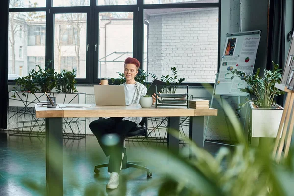 Elegante persona non binaria con caffè per andare e notebook seduto vicino al computer portatile sul posto di lavoro in ufficio — Foto stock