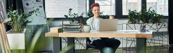 Persona non binaria con caffè da andare e notebook seduto vicino al computer portatile sul posto di lavoro in ufficio, banner — Foto stock