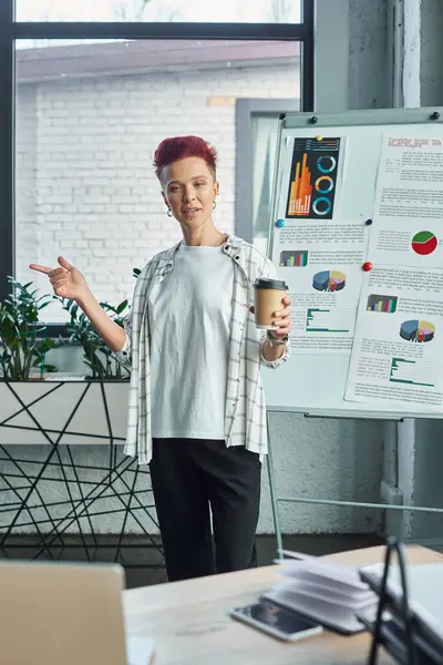 Queer person with coffee to go talking near flip chart with graphs during online call on laptop — Stock Photo