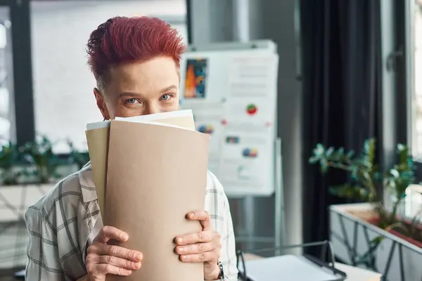 Cheerful bigender person with short hair obscuring face with folders and looking at camera in office — Stock Photo