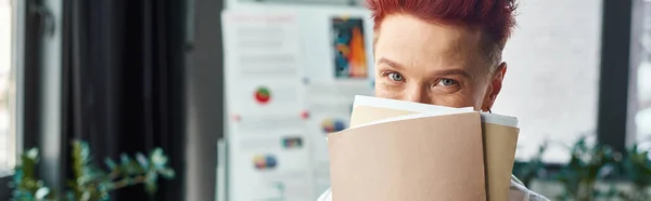 Cheerful bigender person with obscuring face with folders and looking at camera in office, banner — Stock Photo