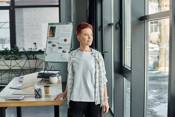 Thoughtful queer person in casual attire standing near windows and looking away in modern office — Stock Photo