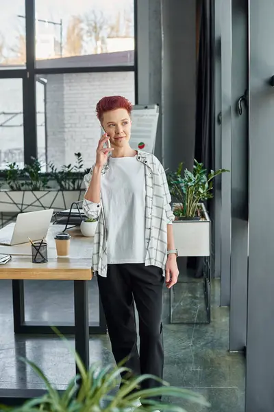 Elegante gerente más grande en atuendo casual de pie y hablando en el teléfono inteligente en la oficina moderna - foto de stock