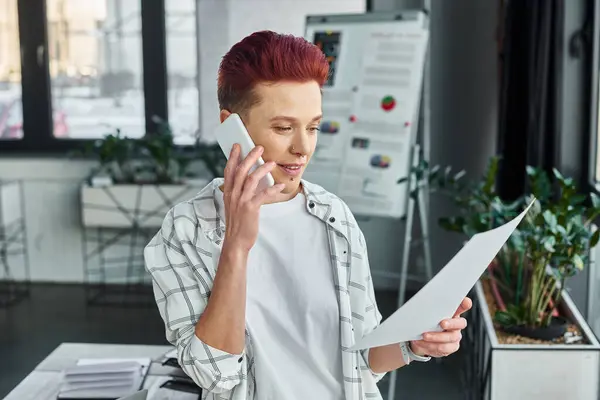 Bigender manager in casual attire looking at document and talking on smartphone in modern office — Stock Photo