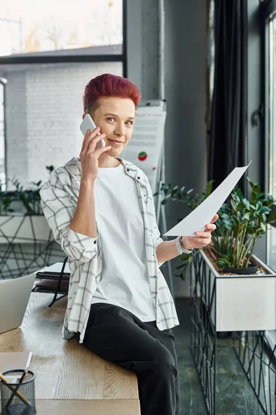 Queer person in casual attire sitting on work desk with document and talking on smartphone in office — Stock Photo