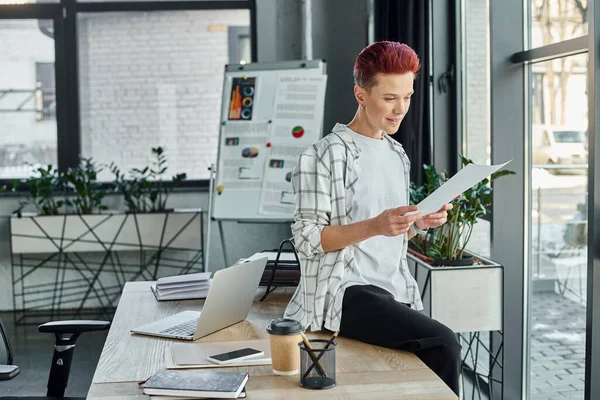 Pessoa não-binário trabalhando com documentos enquanto sentado na mesa de trabalho perto de dispositivos e café para ir — Fotografia de Stock