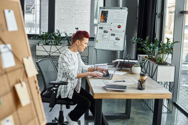 Квір-людина в повсякденному вбранні, що друкується на ноутбуці, сидячи поруч з документами на робочому столі в офісі — стокове фото