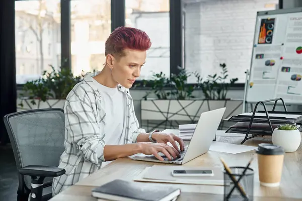 Концентрована квір-людина в повсякденному вбранні, що працює на ноутбуці поблизу кави і смартфона в офісі — стокове фото