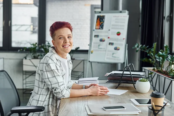 Joyful queer manager sitting at workplace near smartphone and digital tablet and looking at camera — Stock Photo