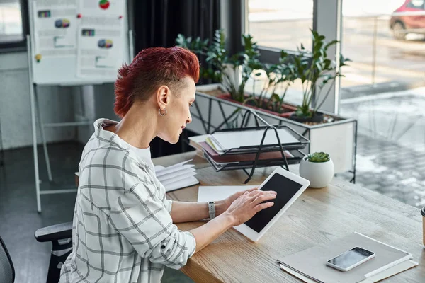 Недвоичный человек в повседневной одежде с помощью цифрового планшета рядом с документами и смартфона в офисе — стоковое фото