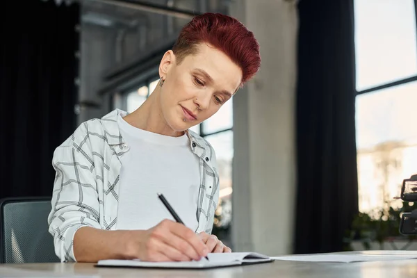 Concentrated stylish bigender person sitting at workplace in modern office and writing in notebook — Stock Photo