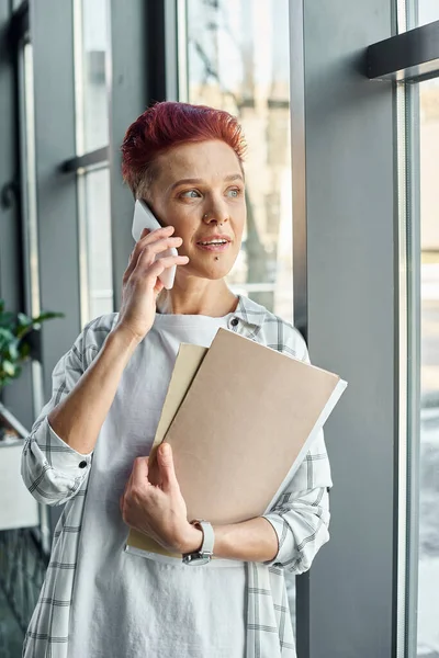 Stylish bigender manager standing near window with documents and talking on smartphone in office — Stock Photo
