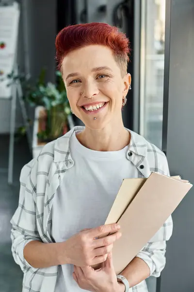 Retrato de alegre queer persona sosteniendo carpeta con documentos y mirando a la cámara en la oficina - foto de stock