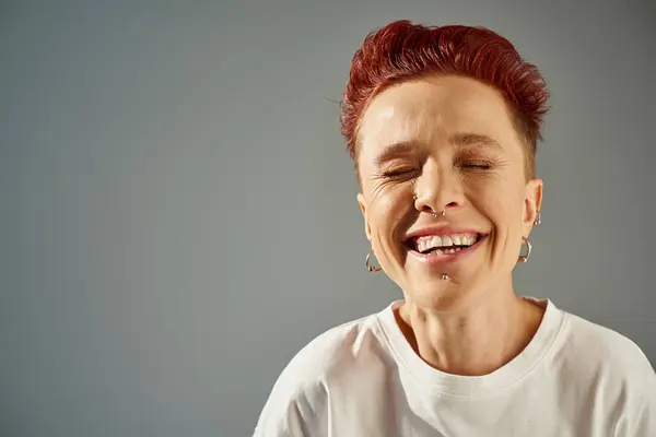 Portrait of redhead bigender person with facial piercing laughing with closed eyes on grey backdrop — Stock Photo