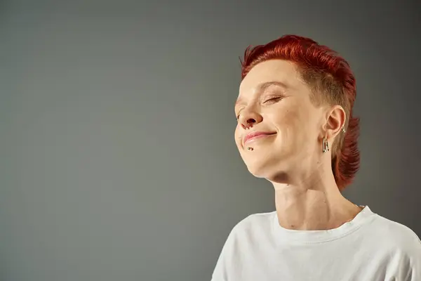 Portrait of redhead bigender person with facial piercing smiling with closed eyes on grey backdrop — Stock Photo