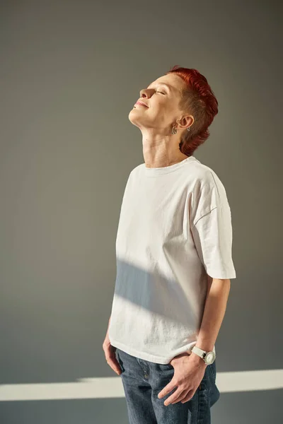 Persona queer feliz y única en camiseta blanca y jeans de pie a la luz del sol sobre fondo gris - foto de stock