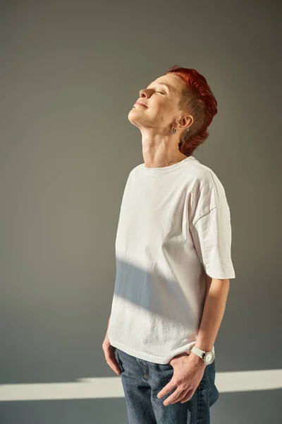 Happy and unique queer person in white t-shirt and jeans standing in sunlight on grey backdrop — Stock Photo