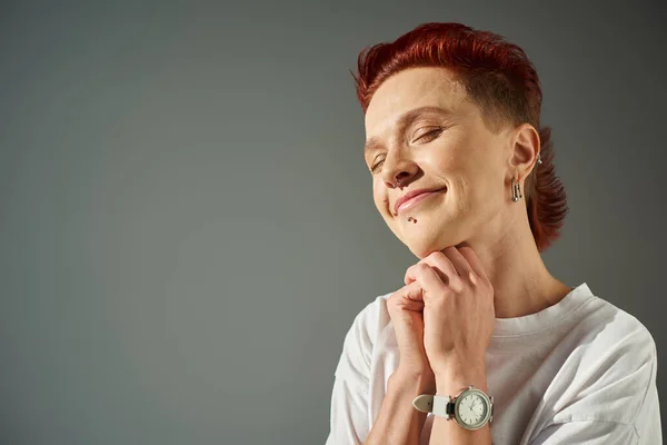 Retrato de pelirroja persona más grande con piercing facial sonriendo con los ojos cerrados sobre fondo gris - foto de stock