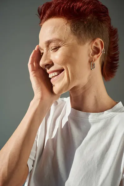 Redhead bigender person with piercing smiling with closed eyes and hand near face on grey, portrait — Stock Photo