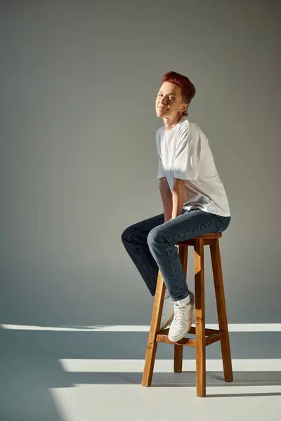 Happy non-binary person in white t-shirt and jeans sitting on stool in sunlight on grey, full length — Stock Photo