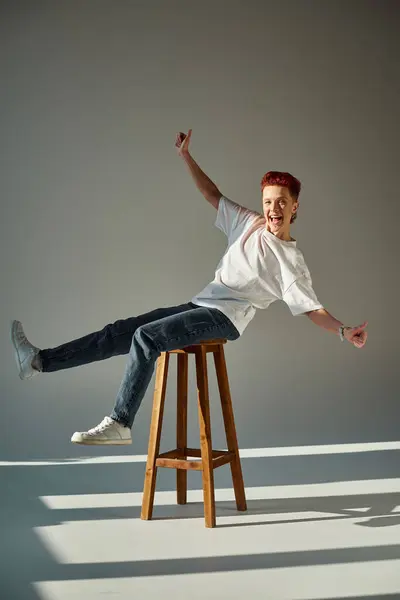 Overjoyed queer person having fun while sitting on stool and showing thumbs up in sunlight on grey — Stock Photo