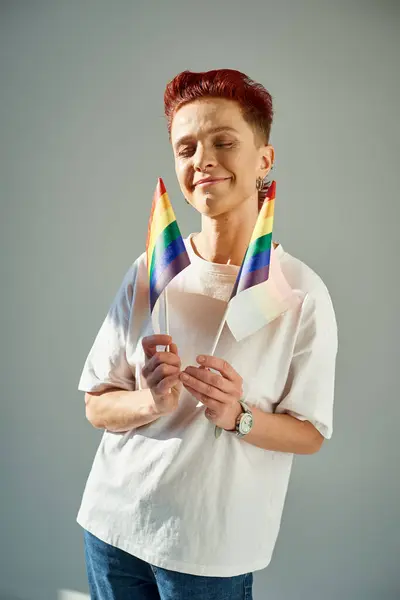 Redhead queer person in white t-shirt holding small LGTB flags and smiling with closed eyes on grey — Stock Photo