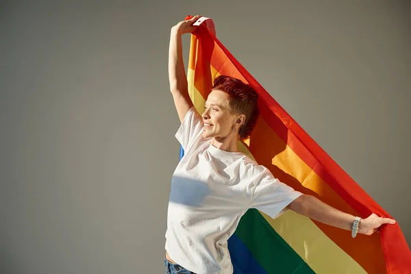 Feliz y única persona queer en camiseta blanca posando con colores arco iris bandera LGBT sobre fondo gris - foto de stock