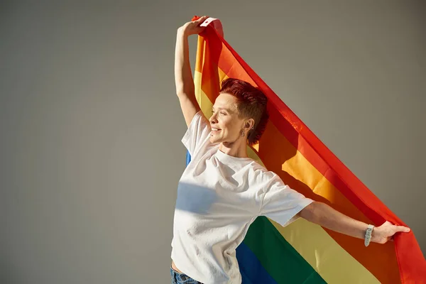 Happy and unique queer person in white t-shirt posing with rainbow colors LGBT flag on grey backdrop — Stock Photo