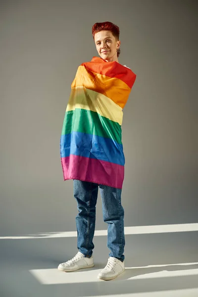 Volle Länge lächelnde queere Person posiert mit Regenbogenfarben LGBT-Flagge weiß auf grau — Stockfoto