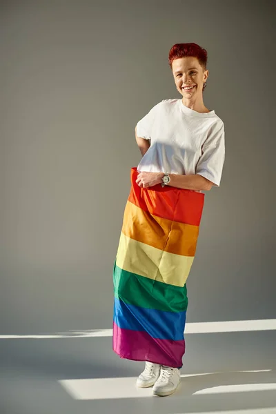 Piena lunghezza di persona queer sorridente in posa con i colori arcobaleno bandiera LGBT bianco in piedi sul grigio — Foto stock