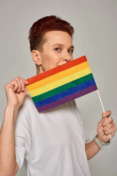 Modello queer rossa in t-shirt bianca in posa con piccolo LGBT piatto vicino alla faccia guardando la fotocamera sul grigio — Foto stock
