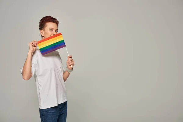 Redhead queer model in white t-shirt posing with small LGBT flat near face looking at camera on grey — Stock Photo